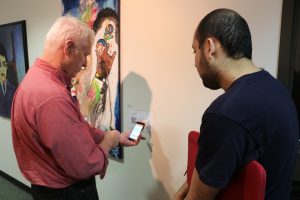 Two men, one with white hair and one with dark hair, looking at a mobile phone in front of artwork